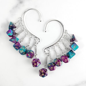 May include: A pair of silver-toned ear cuffs featuring multiple dangling miniature polyhedral dice. Each die displays a gradient of metallic purple and teal colours with gold-coloured numbers. The dice are attached to the cuffs by small silver chains.