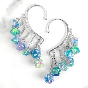 May include: A pair of silver-toned ear cuffs featuring multiple dangling polyhedral dice charms. Each charm is a multi-coloured resin die in shades of blue, green, and purple with white numbers. The dice are attached to the metal cuffs by small silver chains.