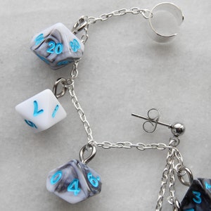 Op de afbeelding: Een zilveren kettingoorring met een zilveren oormanchet en meerdere dobbelsteenbedels. De dobbelstenen zijn zwart-wit met blauwe cijfers. De oorring heeft ook een zilveren oorring met een kleine zilveren bol.
