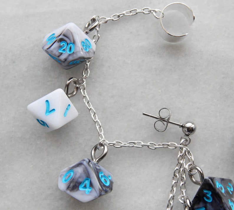 Op de afbeelding: Een zilveren kettingoorring met een zilveren oormanchet en meerdere dobbelsteenbedels. De dobbelstenen zijn zwart-wit met blauwe cijfers. De oorring heeft ook een zilveren oorring met een kleine zilveren bol.