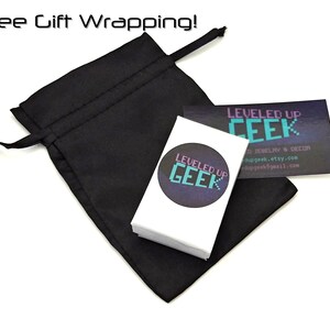 Op de afbeelding: Een zwarte trekkoordtas met een witte kaart erin. De kaart heeft een paars en turquoise logo met de tekst "LEVELED UP GEEK" en bevat de tekst "O JEWELRY & DECOR", "dupgeek.etsy.com" en "dupgeek@gmail.com".