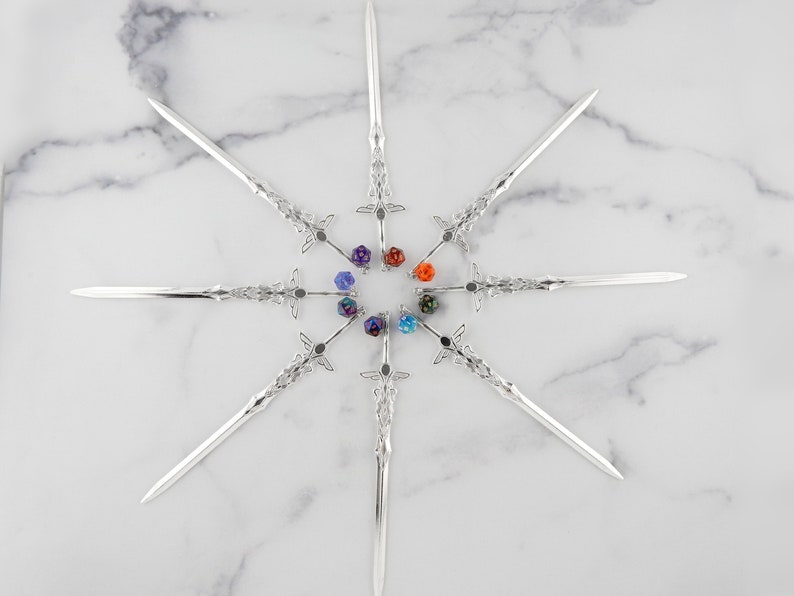 May include: Eight silver sword-shaped hair sticks arranged in a circle around eight colorful acrylic dice. The dice are in shades of purple, blue, green, and orange.