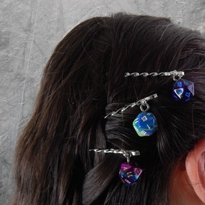 Op de afbeelding: Drie zilveren haarclips met iriserende dobbelstenen bedels. De dobbelstenen zijn blauw, groen en paars met witte cijfers.