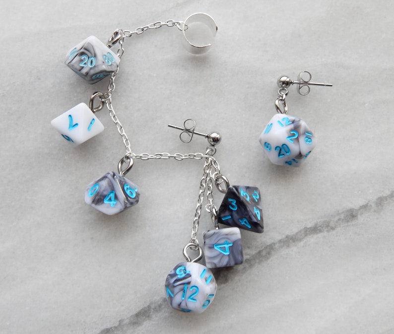 Op de afbeelding: Een paar zilveren oorbellen met een hangende ketting en meerdere witte en grijze dobbelstenen met blauwe nummers. De dobbelstenen zijn in verschillende vormen en maten.