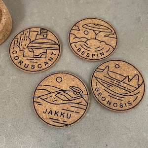 May include: Four round cork coasters with black outlines of different Star Wars locations. The coasters feature the names "Coruscant", "Bespin", "Geonosis", and "Jakku".