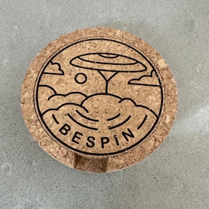 May include: A round cork coaster featuring a black and white line drawing of a tree with a large disc-shaped canopy, clouds, and the word "BESPIN" underneath.