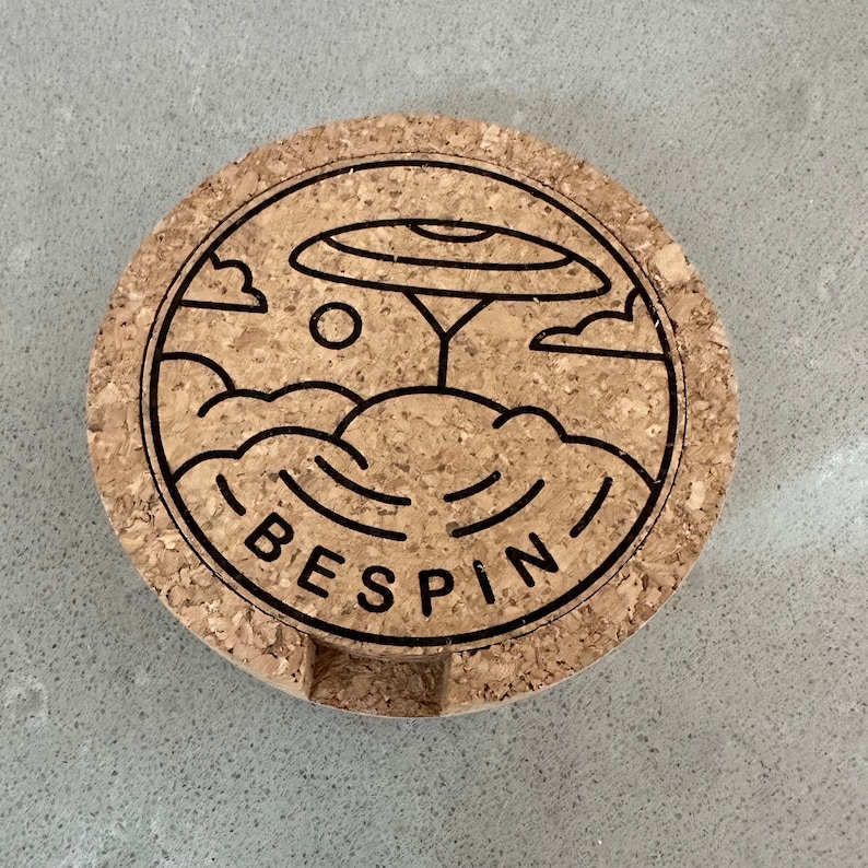 May include: A round cork coaster featuring a black and white line drawing of a tree with a large disc-shaped canopy, clouds, and the word "BESPIN" underneath.