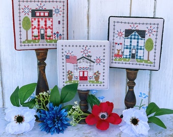 May include: Three framed cross-stitch patterns featuring red, white, and blue houses with fireworks. The houses are decorated with gingham and plaid patterns. The frames are on wooden stands.