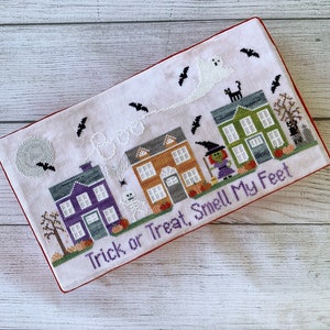 May include: A white cross-stitch piece with a Halloween theme. It features a row of houses with a ghost, witch, and skeleton. The text reads "Boo Trick or Treat, Smell My Feet".