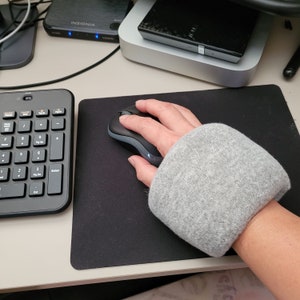 May include: A gray wrist rest with a black computer mouse on a black mouse pad. The mouse is being used by a hand.