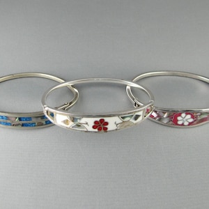 May include: Three silver-toned hinged bangle bracelets. The left bracelet features blue and iridescent inlay. The centre bracelet has a white background with a red flower design. The right bracelet displays a dark red background with a white flower.