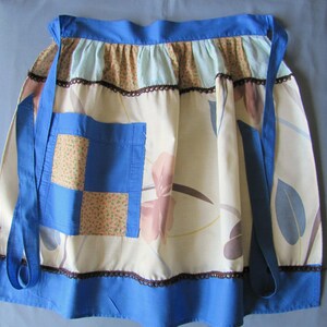 May include: A vintage apron with a cream background and a floral design in pink, blue, and brown. It has a blue pocket and trim, with blue ties. The top edge is ruffled with a contrasting fabric. The apron is designed for kitchen use.