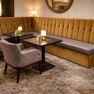 May include: A restaurant seating area with a mustard-coloured velvet booth and a grey leather seat. Two grey armchairs and small black tables with lamps are arranged. The room has a warm, inviting atmosphere, with a large mirror on the wall.