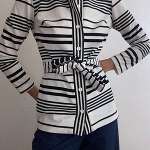 May include: A black and white striped button-down shirt with a matching tie belt. The shirt has long sleeves and a collared neckline. The stripes are horizontal and vary in width. The shirt is paired with dark blue pants.
