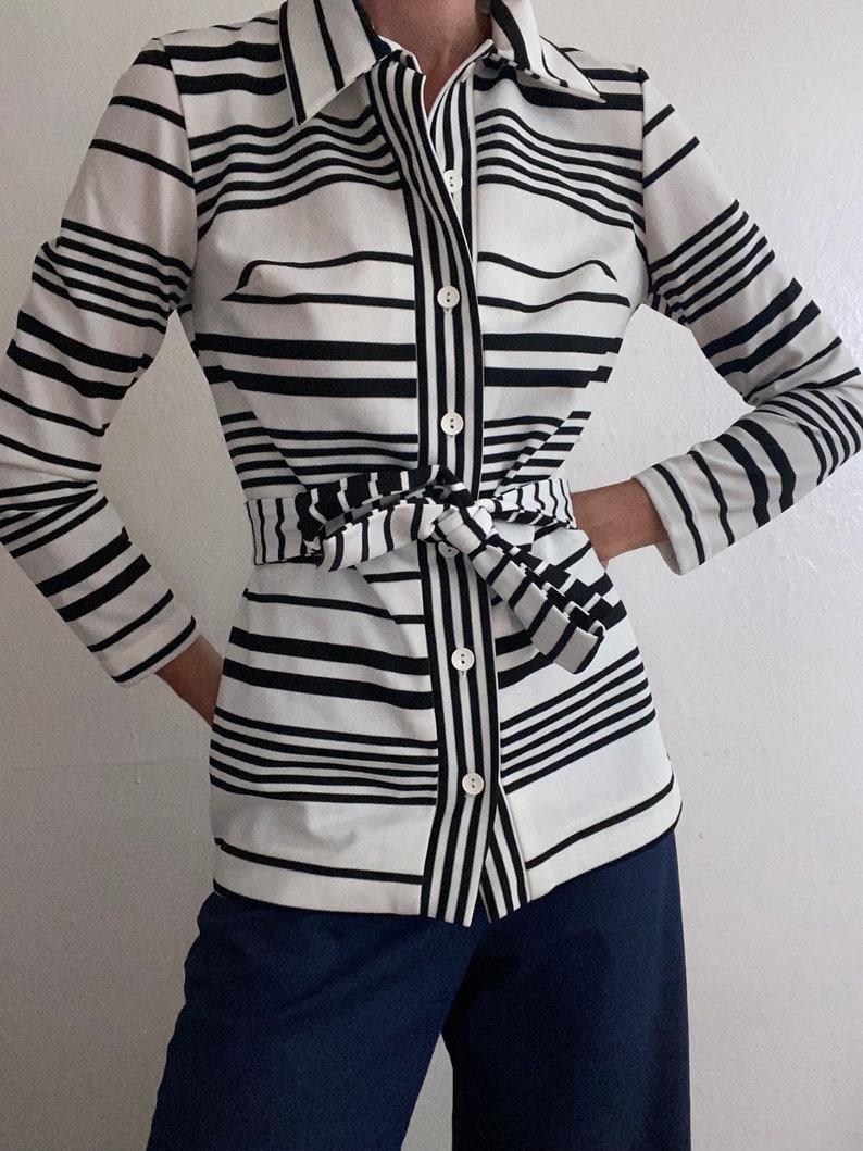 May include: A black and white striped button-down shirt with a matching tie belt. The shirt has long sleeves and a collared neckline. The stripes are horizontal and vary in width. The shirt is paired with dark blue pants.