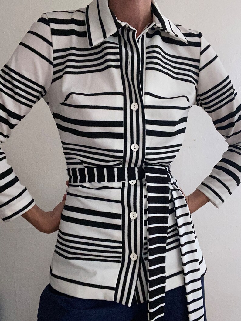 May include: A black and white striped button-down shirt with a matching belt. The shirt features a collared neckline, long sleeves, and a fitted silhouette. The stripes are horizontal and vertical, creating a bold pattern. The belt cinches the waist.