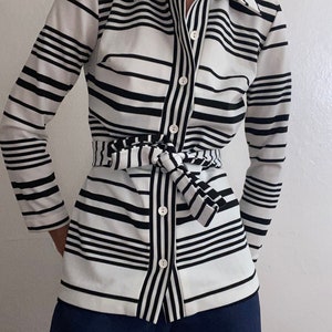 May include: A long-sleeved button-down shirt with a black and white striped pattern. The shirt has a matching belt tied at the waist and is paired with navy blue pants. The shirt features a classic collar and button closure.