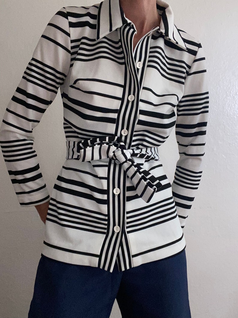 May include: A long-sleeved button-down shirt with a black and white striped pattern. The shirt has a matching belt tied at the waist and is paired with navy blue pants. The shirt features a classic collar and button closure.
