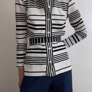 May include: A button-down shirt with a black and white striped pattern. The shirt has long sleeves and a collar. The bottom half of the shirt has a matching black and white striped belt. The pants are a solid navy blue color.