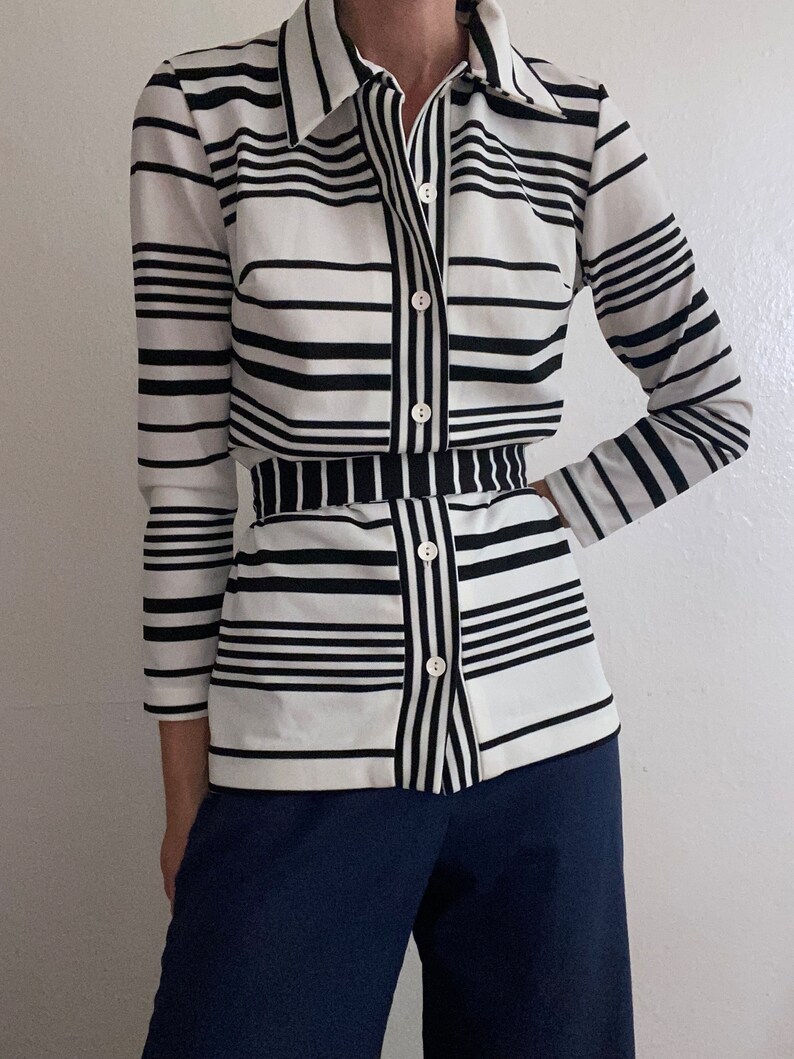 May include: A button-down shirt with a black and white striped pattern. The shirt has long sleeves and a collar. The bottom half of the shirt has a matching black and white striped belt. The pants are a solid navy blue color.