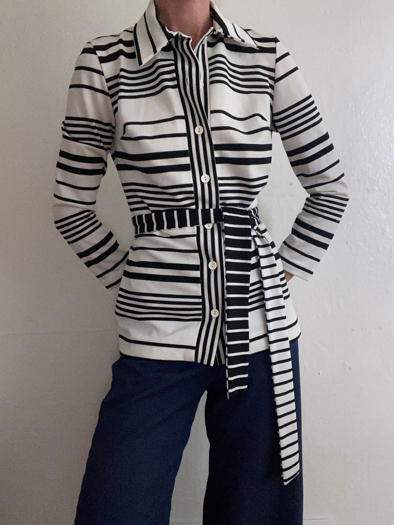 May include: A black and white striped button-down shirt with a matching belt. The shirt has long sleeves and a collared neckline. The model is wearing navy blue pants. The shirt's stripes are horizontal and vertical.