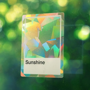 May include: A rectangular sticker featuring a holographic, geometric pattern with shifting colors of green, yellow, and blue. The sticker has a white border and a white rectangular section at the bottom containing the black printed text Sunshine.