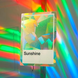 May include: A rectangular card featuring a holographic, multi-faceted geometric pattern in shades of green, yellow, and blue. The lower portion contains a white rectangular block with the word Sunshine printed in bold, black, sans-serif text.
