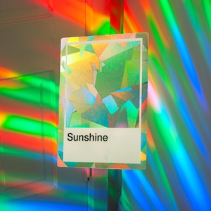 May include: A rectangular card with the word "Sunshine" on a white label. The card has a geometric design with iridescent colors. The background is filled with colorful light reflections in shades of red, green, and blue.