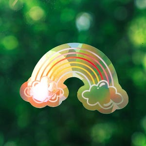 May include: A holographic rainbow sticker featuring a multi-colored arched design with a cloud at each end. The sticker has a reflective, prismatic surface that catches light, displaying various colors across its curved bands and cloud shapes.