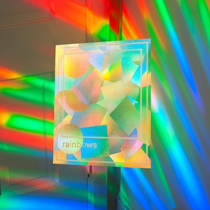 May include: A square, iridescent artwork with the word "rainbows" in a light font. The artwork is illuminated by colorful light beams, creating a vibrant display of colors. The artwork features geometric shapes in various colors.