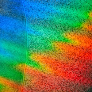 May include: Close-up of a colorful, textured fabric with a rainbow gradient. The colors transition from blue to green, yellow, orange, and red. A vertical seam runs down the left side of the image.
