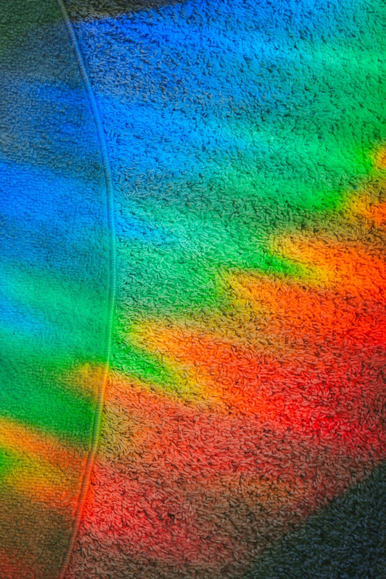 May include: Close-up of a colorful, textured fabric with a rainbow gradient. The colors transition from blue to green, yellow, orange, and red. A vertical seam runs down the left side of the image.