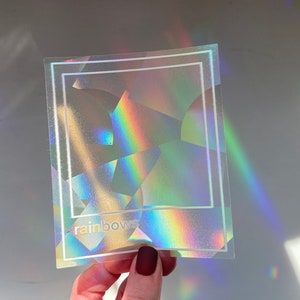 May include: A holographic sticker with a geometric design and the word "rainbows" in white. The sticker reflects a spectrum of colors, including red, orange, yellow, green, blue, and purple. A white border frames the design.