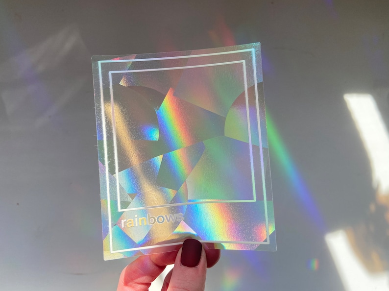 May include: A holographic sticker with a geometric design and the word "rainbows" in white. The sticker reflects a spectrum of colors, including red, orange, yellow, green, blue, and purple. A white border frames the design.