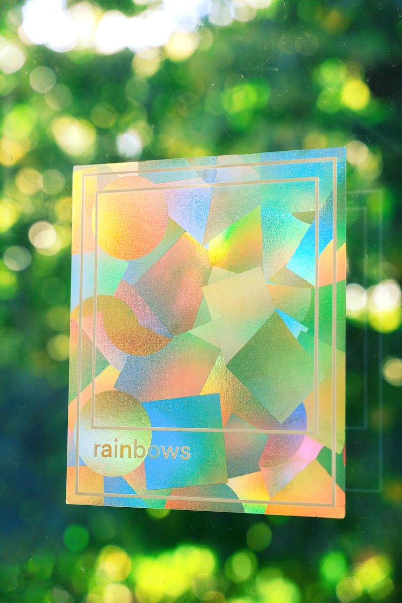 May include: A holographic sticker with a geometric design of squares, circles, and other shapes in shades of orange, green, blue, and gold. The word "rainbows" is printed in a light color at the bottom.