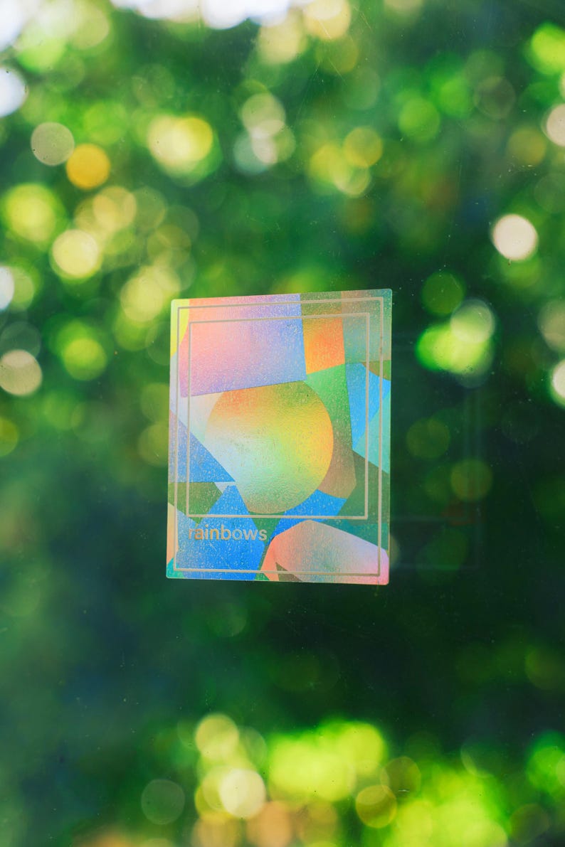 May include: A rectangular sticker with a colorful abstract design featuring a rainbow gradient and geometric shapes. The word "rainbows" is printed at the bottom. The sticker is set against a blurred green background.