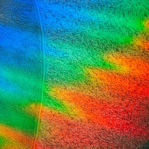 May include: A textured, soft fabric surface featuring a vibrant, prismatic light refraction pattern. The light displays a spectrum of colors including red, orange, yellow, green, and blue, casting a rainbow effect across the material's fibers.