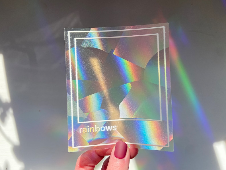 May include: A holographic sticker with a white border and the word "rainbows" at the bottom. The sticker has a geometric design that reflects light, creating a rainbow effect. The sticker is held by a person.