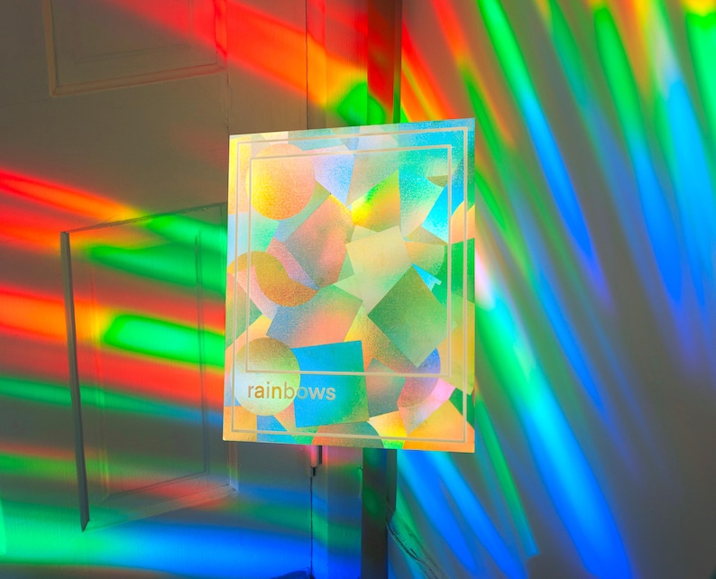 May include: A square, iridescent artwork with the word "rainbows" in a light font. The artwork is illuminated by colorful light beams, creating a vibrant display of colors. The artwork features geometric shapes in various colors.