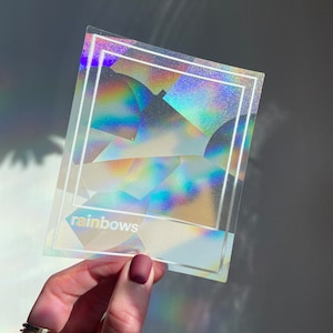 May include: A clear, holographic sticker with a rainbow effect, featuring a white border and the word "rainbows" at the bottom. The sticker is held in a hand with a gold ring and a matte burgundy nail polish.