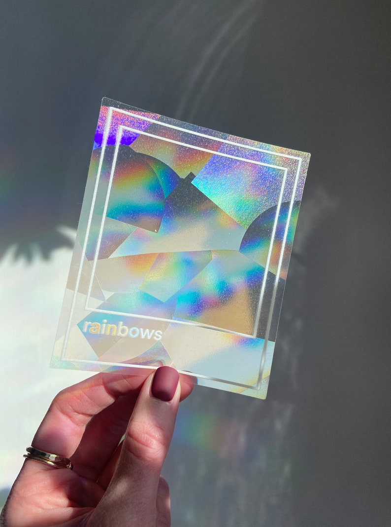 May include: A clear, holographic sticker with a rainbow effect, featuring a white border and the word "rainbows" at the bottom. The sticker is held in a hand with a gold ring and a matte burgundy nail polish.