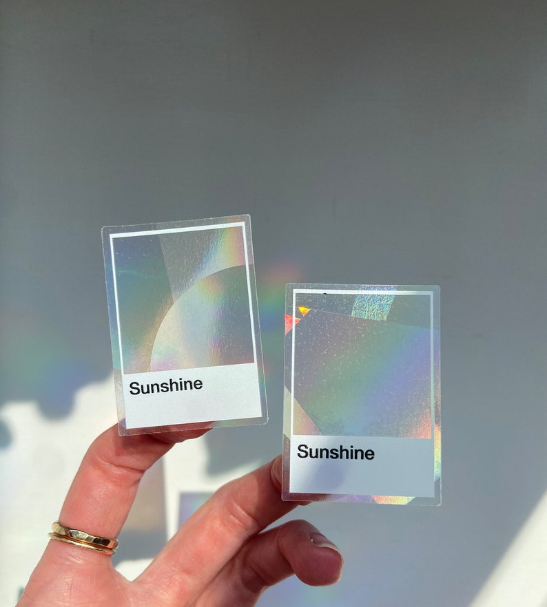 May include: Two rectangular stickers featuring a holographic finish that reflects rainbow colors. Each sticker has a white border and a white rectangular section at the bottom with the word Sunshine printed in black sans-serif font.