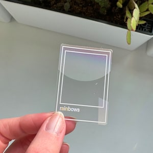 May include: A clear, rectangular sticker with a white border and the word "rainbows" in a lowercase, sans-serif font. The sticker has a holographic effect, reflecting a spectrum of colors. The sticker is held by a person's hand.