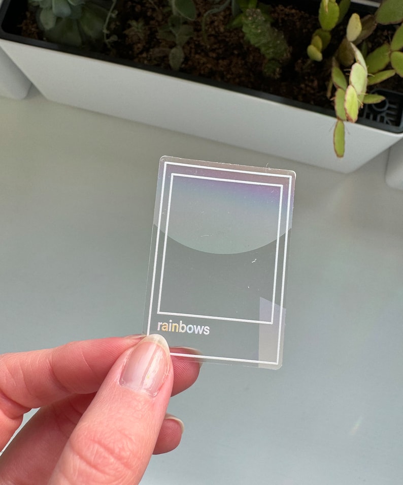 May include: A clear, rectangular sticker with a white border and the word "rainbows" in a lowercase, sans-serif font. The sticker has a holographic effect, reflecting a spectrum of colors. The sticker is held by a person's hand.