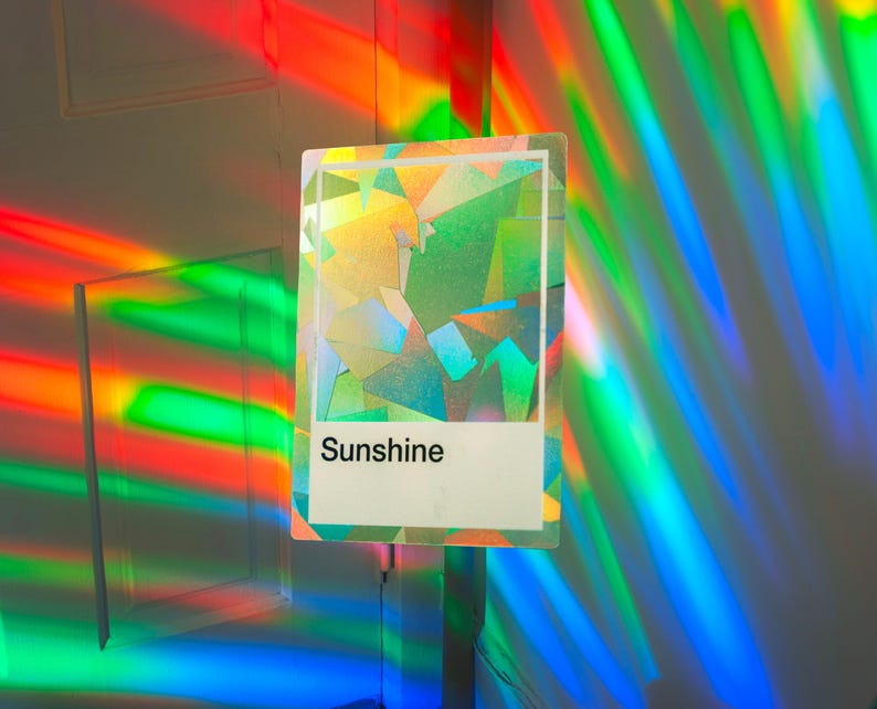 May include: A rectangular sticker featuring a holographic, geometric, multi-colored pattern with a white rectangular block at the bottom containing the black text Sunshine. The sticker reflects vibrant rainbow light beams across the surrounding area.