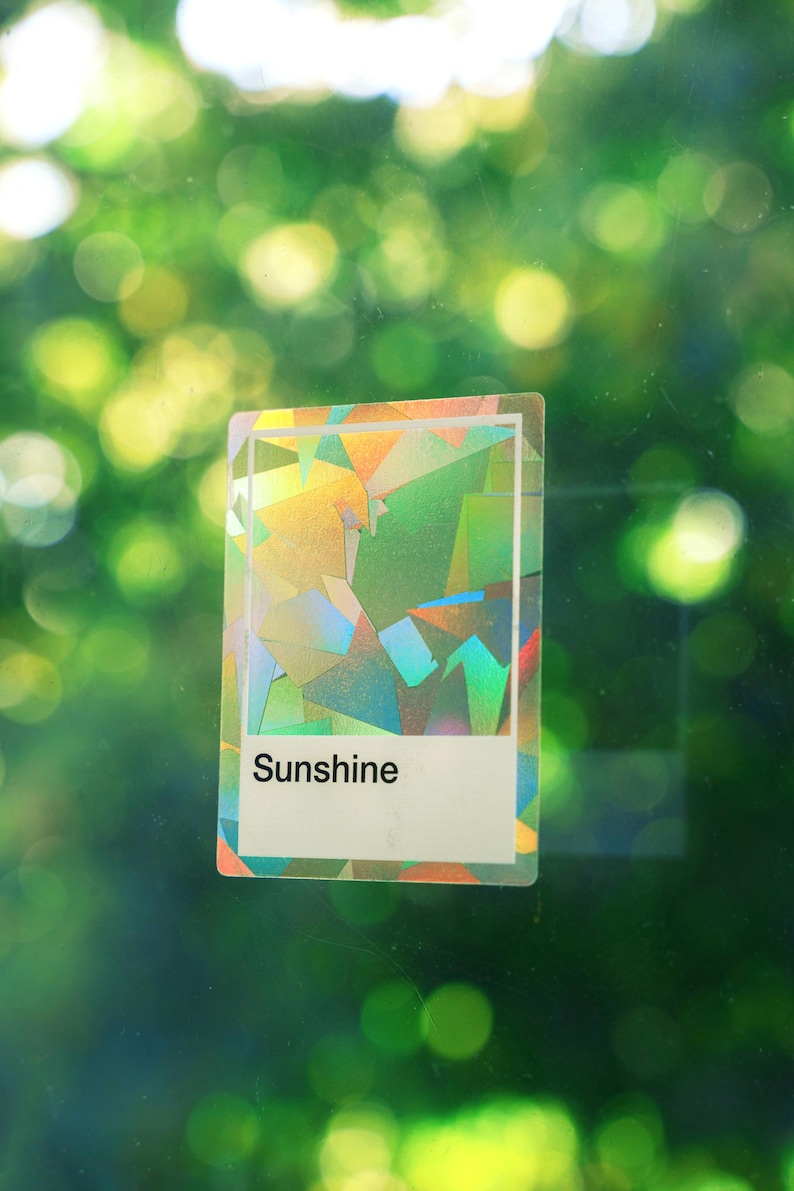 May include: A rectangular sticker featuring a holographic, geometric pattern with shifting colors of green, yellow, and blue. The sticker has a white border and a white rectangular section at the bottom containing the black printed text Sunshine.