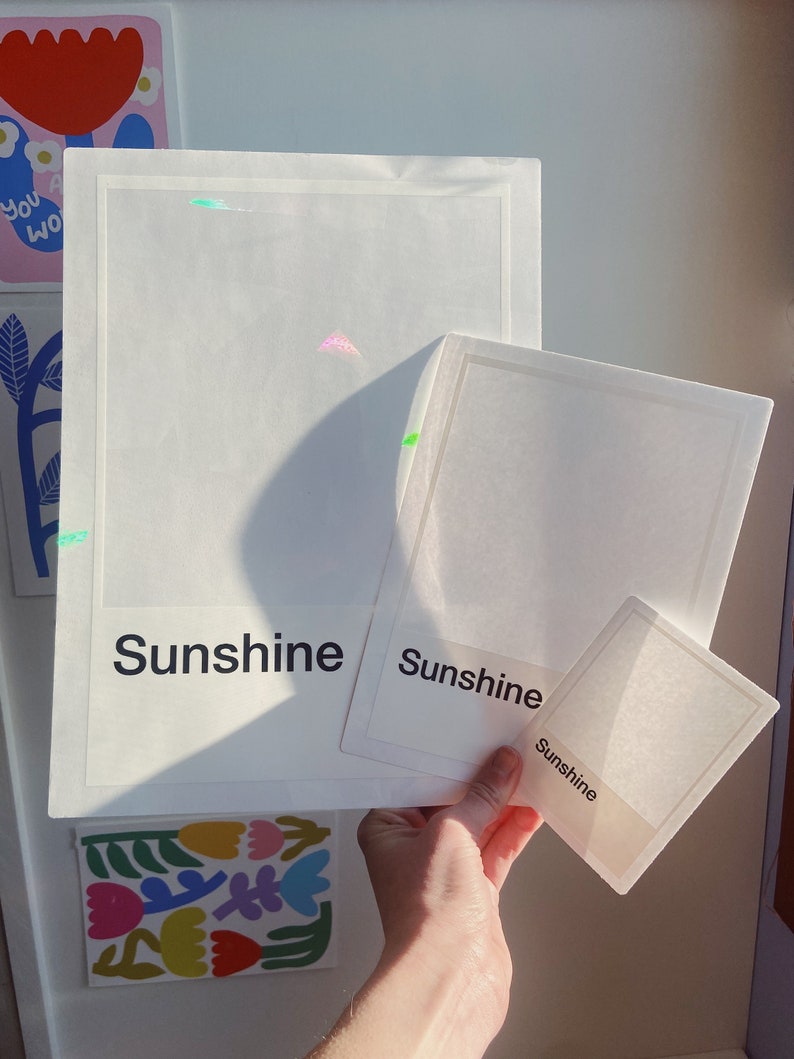 May include: Three rectangular white cards of varying sizes, each featuring a thin border and the word "Sunshine" printed in black text at the bottom. The cards are semi-transparent, allowing light to pass through and cast shadows.