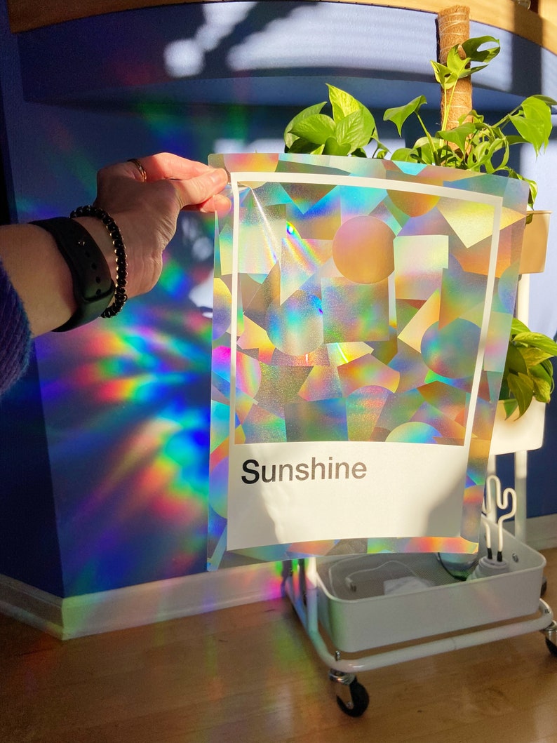 May include: A holographic sticker with a geometric pattern and the word "Sunshine" printed on it. The sticker is iridescent and reflects light in a rainbow of colors.
