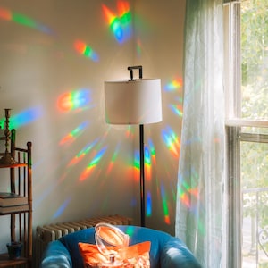 May include: A floor lamp with a white lampshade and a black metal frame. The light from the lamp is refracted into a rainbow of colors on the wall. A blue armchair with an orange pillow is in the foreground.