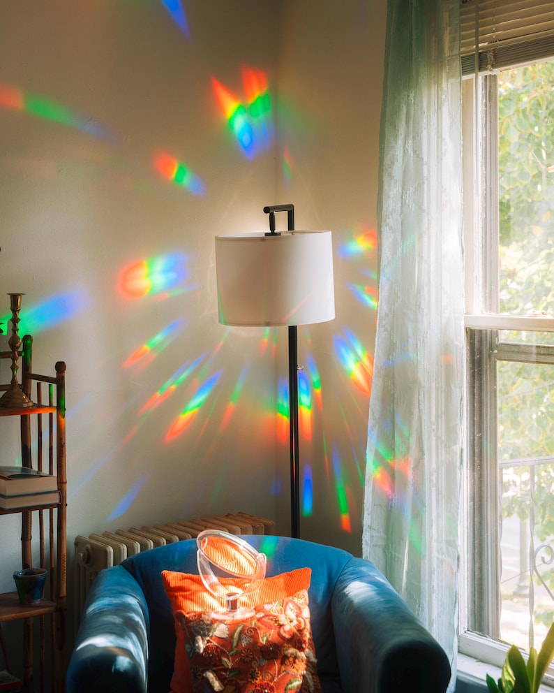 May include: A floor lamp with a white lampshade and a black metal frame. The light from the lamp is refracted into a rainbow of colors on the wall. A blue armchair with an orange pillow is in the foreground.